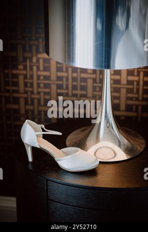 Chaussure blanche de mariée sur la table sous la lampe moderne, élégant détail de mariage Banque D'Images