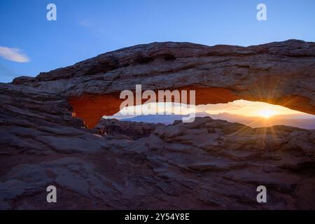 Vue imprenable sur le lever du soleil à Mesa Arch dans le parc national de Canyonlands, Utah. Les rayons du soleil illuminent l’arche et le paysage accidenté. Banque D'Images