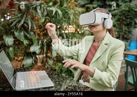 Femme assise à la table de graphite avec ordinateur portable et gesticulant dans l'air tout en jouant au jeu VR Banque D'Images