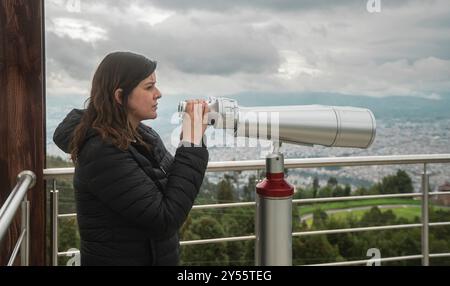 Vue de côté d'une belle jeune femme, debout à une plate-forme d'observation, tenant dans ses mains de longues jumelles montées sur un piédestal métallique contre un nuageux Banque D'Images