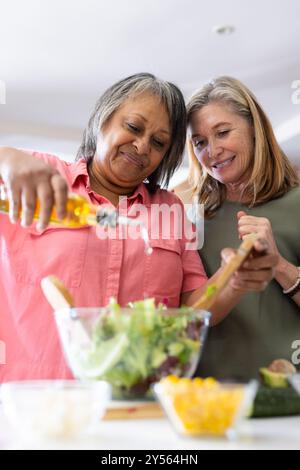 À la maison, la préparation de salade, deux amies féminines multiraciales seniors ajoutant des ingrédients Banque D'Images