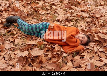 garçon latino regardant la caméra, allongé sur le sol plein de feuilles d'automne tombées. Banque D'Images