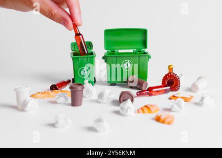 Scène miniature représentant la main collectant les ordures dispersées dans des bacs de recyclage verts sur un fond blanc propre. Banque D'Images