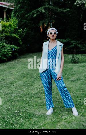 Une femme à la mode se tient confiante sur une pelouse verte, portant une combinaison bleue audacieuse à motifs géométriques, un gilet sans manches léger et des baskets blanches Banque D'Images