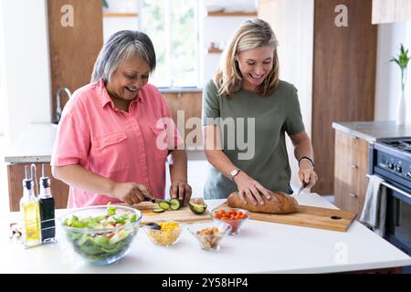 À la maison, préparant le repas, deux amies multiraciales séniors coupant des légumes et du pain Banque D'Images