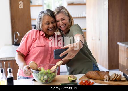 À la maison, prenant selfie, deux amies féminines multiraciales seniors souriant et préparant de la salade en cuisine Banque D'Images