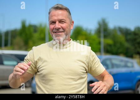 Un homme mûr joyeux portant un t-shirt jaune clair, se déplaçant de façon décontractée à l'extérieur par une journée ensoleillée, avec des voitures et de la verdure en arrière-plan. Banque D'Images