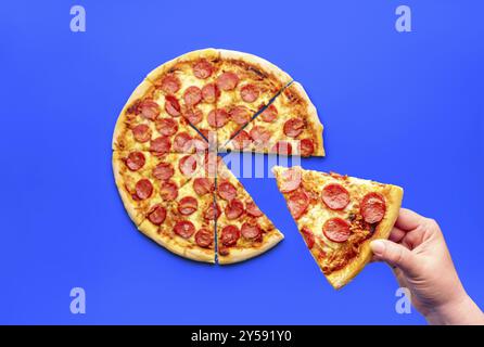 Main de femme prenant une tranche de pizza, vue au-dessus. Délicieuse pizza au pepperoni isolée sur fond bleu Banque D'Images