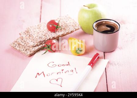 Collation santé avec des biscottes de blé entier, de légumes, de pomme, une tasse de café et une serviette avec un bon matin message sur un fond rose, dans daylig Banque D'Images