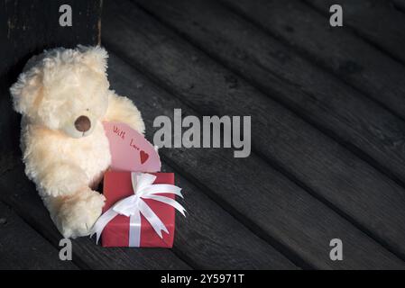 Boîte cadeau rouge avec blanc attaché arc et un adorable ours en peluche jouet tenant un message sur un plancher en bois rustique Banque D'Images