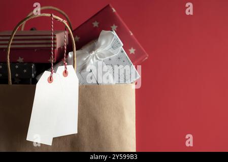 Deux étiquettes blanches non écrites suspendues à un sac en papier rempli de boîtes cadeaux colorées, sur un fond rouge Banque D'Images