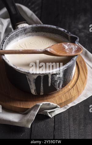 Lait bouilli dans une casserole noire et renversé sur les bords, une cuillère en bois, sur une planche à découper, sur une table rustique. Image en basse lumière Banque D'Images