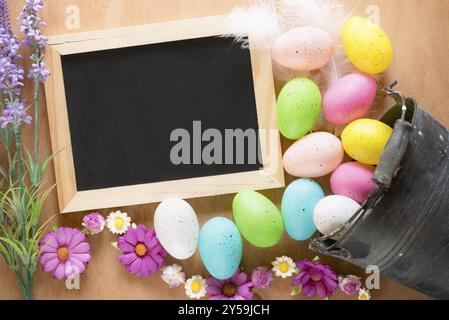 Carte de Pâques avec un seau en métal rustique plein d'œufs peints colorés, renversé sur la table et un tableau noir non écrit entouré de fleurs Banque D'Images