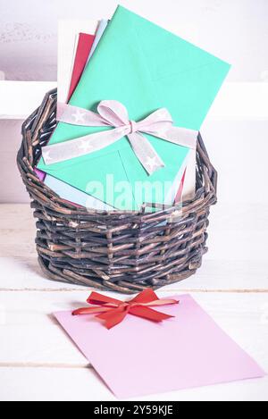 Panier en osier plein de lettres multicolores liées ensemble avec un ruban et une carte de message vierge avec un noeud rouge, sur une table en bois blanc Banque D'Images