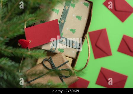 Vue de dessus d'un sac en papier rempli de boîtes cadeaux, sous un sapin de Noël, avec une note en papier rouge vierge clippée sur les poignées et les enveloppes. Tradition de Noël Banque D'Images