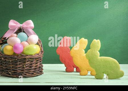Conception de bannière de Pâques avec un panier en osier plein d'œufs peints colorés, avec un noeud en ruban et trois biscuits en forme de lapin, sur un fond vert Banque D'Images