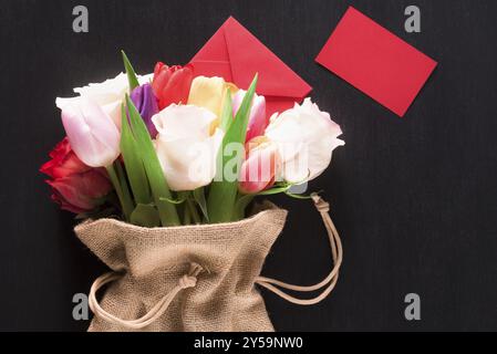 Carte de voeux avec un bouquet de roses et tulipes, enveloppé dans un sac de hesse, une enveloppe fermée et une note de papier rouge, sur un fond de bois noir Banque D'Images