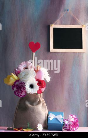 Carte de fête des mères avec des fleurs colorées et une forme de coeur rouge dans un sac de hesse, une boîte cadeau bleue sur une table rose et un tableau noir sur le mur violet Banque D'Images