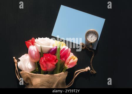 Bouquet coloré de fleurs diverses dans un sac en jute, une montre de poche vintage et une carte de message bleue vierge, sur fond noir Banque D'Images