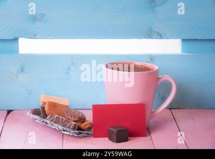 Tasse de café près d'une pile de bonbons sur une écorce d'arbre et une note de papier rouge vierge, sur une table en bois rose et une clôture bleue en arrière-plan Banque D'Images