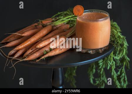 Concept végétarien et diététique avec un verre de jus de carotte frais et un bouquet de carottes fraîchement récoltées, sur une table ronde noire, en basse lumière Banque D'Images