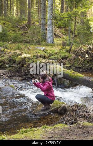 Jeune belle femme randonneur prenant des photos avec son téléphone tout en étant assis sur un rocher au milieu d'un ruisseau de montagne dans la Forêt Noire, Allemagne, Europe Banque D'Images