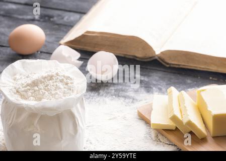 Image de thème de cuisson avec un sac de farine de blé, beurre et œufs, sur une table en bois noir, et un livre ouvert en arrière-plan Banque D'Images