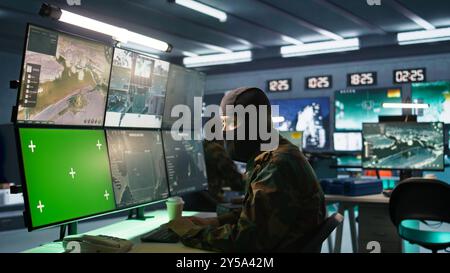 Agent masqué du corps de renseignement supervisant les opérations de sécurité nationale avec un équipement maquette. Espion des forces spéciales examinant les données dans la salle de surveillance avec des moniteurs chroma Key, caméra B. Banque D'Images