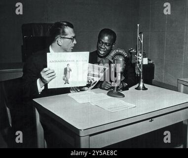 Français : Louis Armstrong (Satchmo) dans l'atelier de Willis Conover. Un microphone et une trompette sont sur la table devant eux. Conover détient un exemplaire de l'album Ambassador Satch d'Armstrong. Banque D'Images