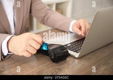 Homme effectuant le paiement via le terminal tout en utilisant l'ordinateur portable à la table en bois, gros plan Banque D'Images