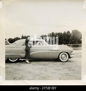 Une femme souriante se penche contre une voiture Buick vintage des années 1950 élégante, profitant d'un moment de légèreté tandis que le conducteur regarde. Le design épuré de l'automobile classique et le cadre extérieur décontracté évoquent l'esprit insouciant des Road trips du milieu du siècle. Buick des années 1950, reconnaissable à sa garniture latérale caractéristique « Sweepspear », sa carrosserie arrondie et ses prises d'air sur l'aile. Les modèles Buick de cette époque, comme le Buick Super ou le Buick Roadmaster, avaient ces caractéristiques de conception distinctes, en particulier les détails chromés sur les côtés. Banque D'Images