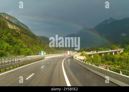 Une autoroute sinueuse traverse des montagnes verdoyantes, où les gouttes de pluie brillent sous la lumière pâle du soleil. Un arc-en-ciel éclatant arque magnifiquement, ajoutant de la couleur Banque D'Images