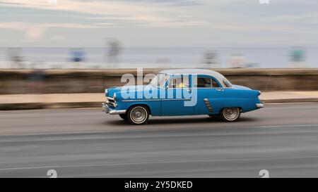 738 vieille voiture classique américaine bleue au toit blanc -Ford de 1952- conduite sur la promenade Malecon, les habitants de la région sur le parapet du front de mer. La Havane-Cuba. Banque D'Images