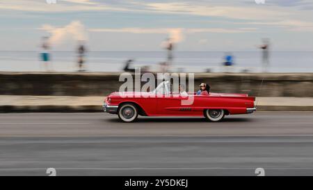 740 vieille voiture classique américaine rouge cramoisi -Ford de 1959- conduite sur la promenade Malecon avec les habitants locaux sur le parapet du front de mer. La Havane-Cuba. Banque D'Images