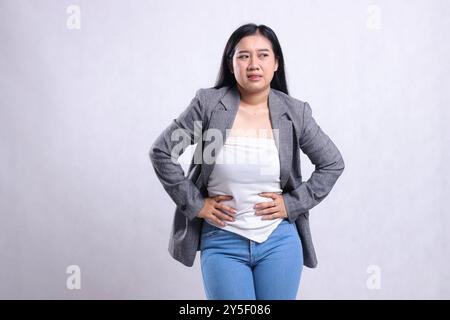 geste belle jeune femme joyeuse indonésie froncant les sourcils maux d'estomac candide portant costume gris et jeans debout isolé sur fond blanc. pour lui Banque D'Images