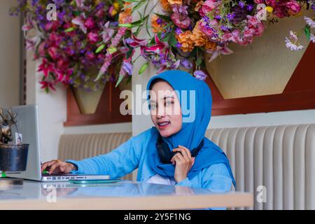 portrait de belle jeune femme adulte portant foulard souriant à la caméra tapant des données dans l'ordinateur portable tout en tenant le casque autour du cou dans le cof intérieur Banque D'Images