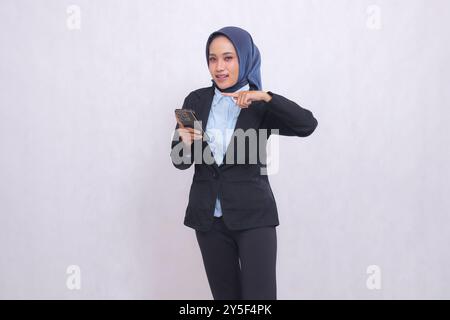 Jeune femme de bureau indonésienne portant un hijab se tient heureux devant la caméra et pointe vers la droite tout en transportant un gadget de téléphone portable. Belle musulmane Banque D'Images