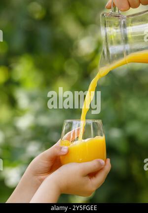 mains d'enfant tenant le verre et le jus d'orange coulant de la cruche avec jardin vert sur le fond Banque D'Images