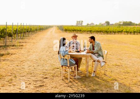 Trois amis se réunissent autour d'une table rustique dans un vignoble, dégustant du vin, des fruits frais, et la compagnie de l'autre sur une journée ensoleillée remplie de joie et de lau Banque D'Images