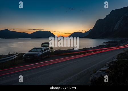Nuit de camping de voiture pendant la nuit chaude coucher de soleil nord sur la plage de mer rocheuse avec des montagnes et des sentiers de feux de circulation, Lofoten Norvège Banque D'Images