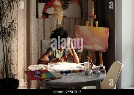 Peintre travaillant sur toile dans un studio lumineux avec diverses fournitures d'art et peintures autour. Lumière diffusée à travers la fenêtre, ajoutant de l'éclairage à l'espace créatif Banque D'Images