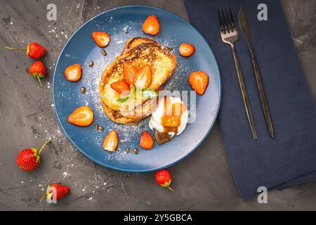 Pain grillé garni de fraises et de feuilles de menthe avec de la crème glacée arrosée de sauce caramel et de sucre en poudre sur une assiette bleue accompagnée de Banque D'Images