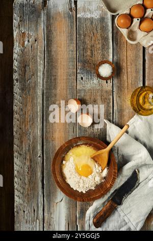 Vue de dessus à un bol rempli de farine sur une table en bois rustique, avec un oeuf cassé ouvert au centre. A proximité se trouvent des œufs supplémentaires et une bouteille de Banque D'Images