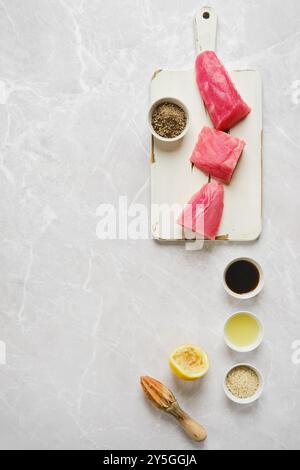 Vue de dessus de filets de thon fraîchement coupés disposés sur une planche à découper blanche avec divers assaisonnements et un citron pressé, prêt pour la préparation i. Banque D'Images