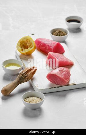 Les steaks de thon frais sont disposés sur une planche à découper à côté du citron pressé, de l'huile d'olive et de petits bols de diverses épices, prêts à être préparés dans un k Banque D'Images