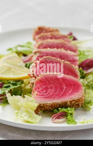 De tendres morceaux de thon poêlé sont tranchés et disposés soigneusement sur une assiette, accompagnés de verts croquants et de quartiers de citron. Les couleurs vives du thon an Banque D'Images