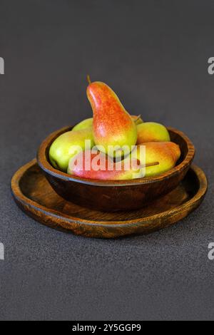 Une collection de poires mûres de différentes formes de rouge et de vert remplit un bol en bois rustique placé sur un fond sombre. Ce charmant arrangement ajoute Banque D'Images