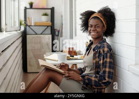 Artiste féminine souriante travaillant sur un projet d'art dans un espace studio lumineux avec diverses fournitures de peinture autour, appréciant le processus créatif Banque D'Images