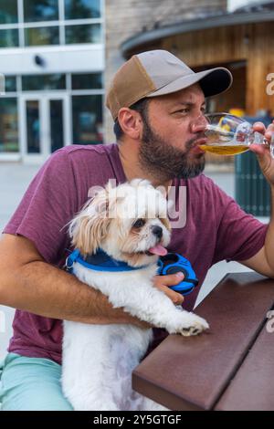 Un homme est assis à une table avec un chien et boit un verre de bière. Le chien porte un collier bleu et il apprécie l'attention. La scène est c. Banque D'Images