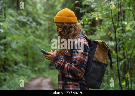 Femme dans un bonnet jaune et chemise à carreaux vérifie son smartphone tout en marchant à travers une forêt verdoyante, portant un sac à dos. Banque D'Images